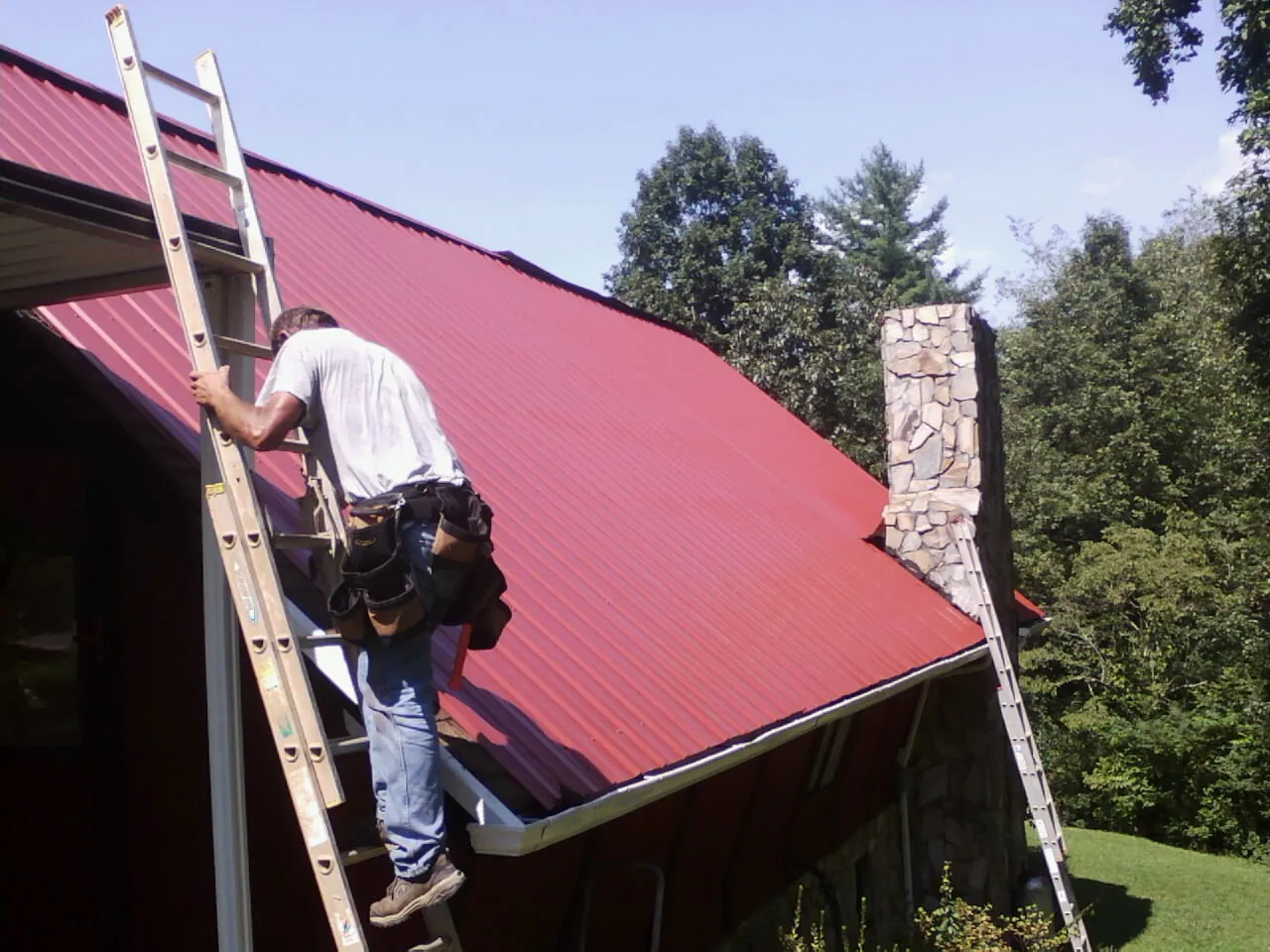 Roofer on a ladder beside a red metal roof and stone chimney — Soffit & Fascia Repair by Roofing Brothers in South Deerfield